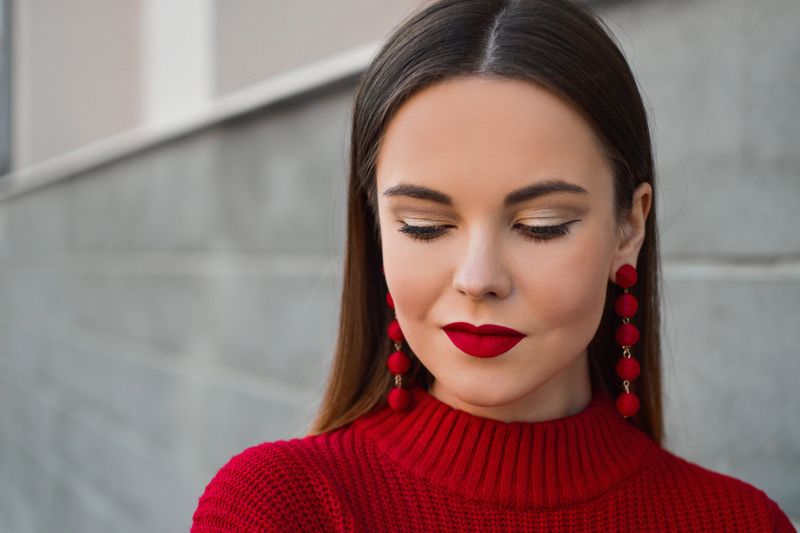 A model wearing red lipstick, red earrings, and a red sweater, posing in front of a brick wall.