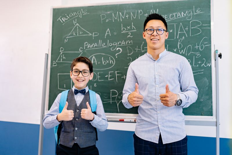 teacher and student in front of green blackboard, both giving the double thumbs up sign