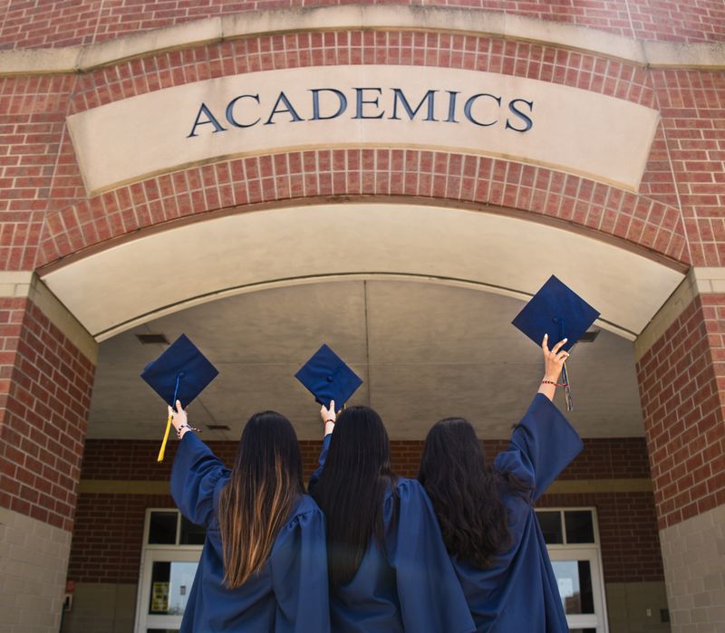 Three people wearing graduation gowns are holding their graduation caps up towards a brick building labeled academics.  