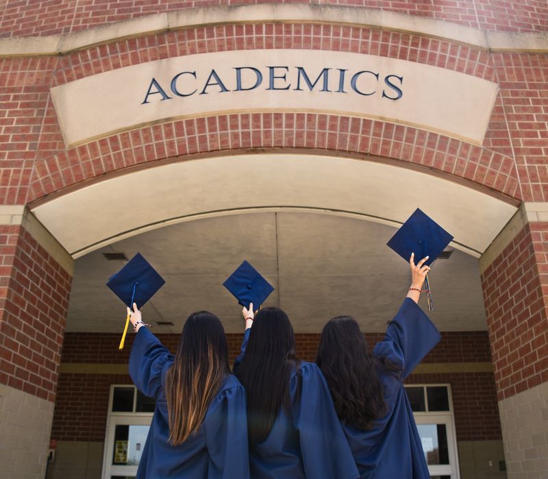 Three people wearing graduation gowns are holding their graduation caps up towards a brick building labeled academics.