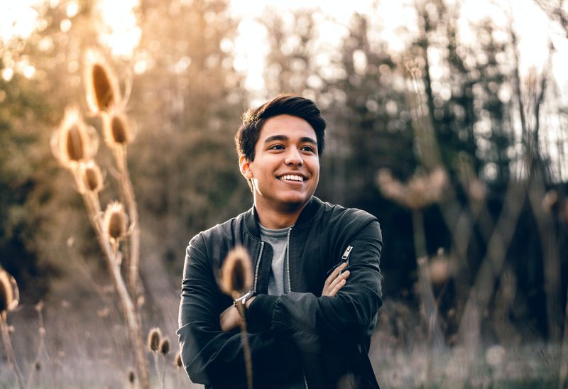 Young smiling man stands in sunlit field with arms crossed while wearing a jack.