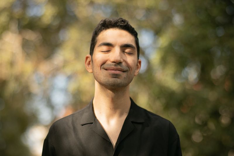 A man in a black shirt, surrounded by green trees, smiling with his eye closed. 