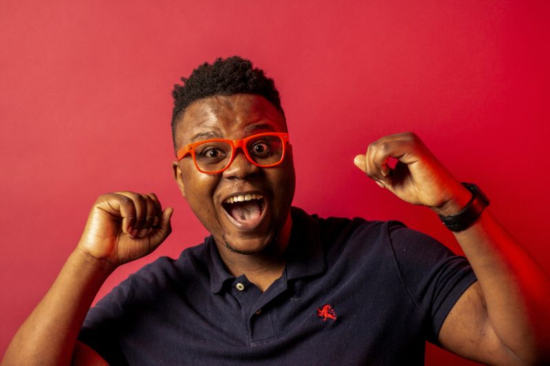 A black man with short afro haircut and orange glasses raises his hands in the air with a joyful expression.