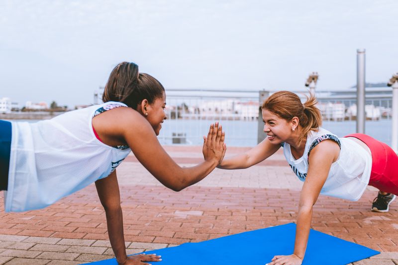 Two women at an outdoor gym giving each other a high five while planking.