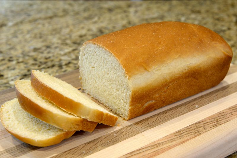 A loaf of white bread with 3 pieces cut off laying on a wooden board on a granite countertop.