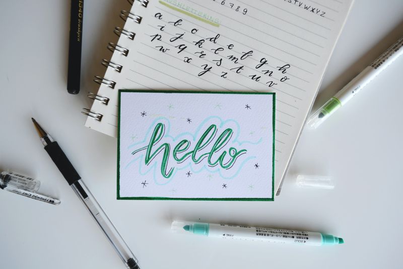 A photo of a stock card saying Hello in lower case written in green ink with a notebook and pens in the background.