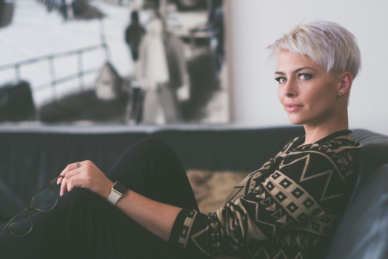 A woman with a short, blonde haircut holds her glasses and casually reclines on a couch in an office. 