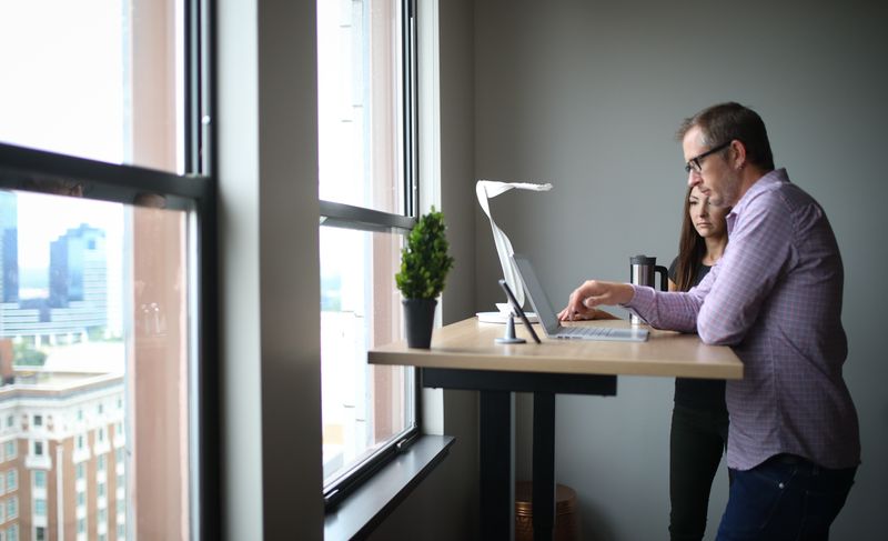 A man and woman standing at a standing desk. The woman watches as the man works on the laptop.