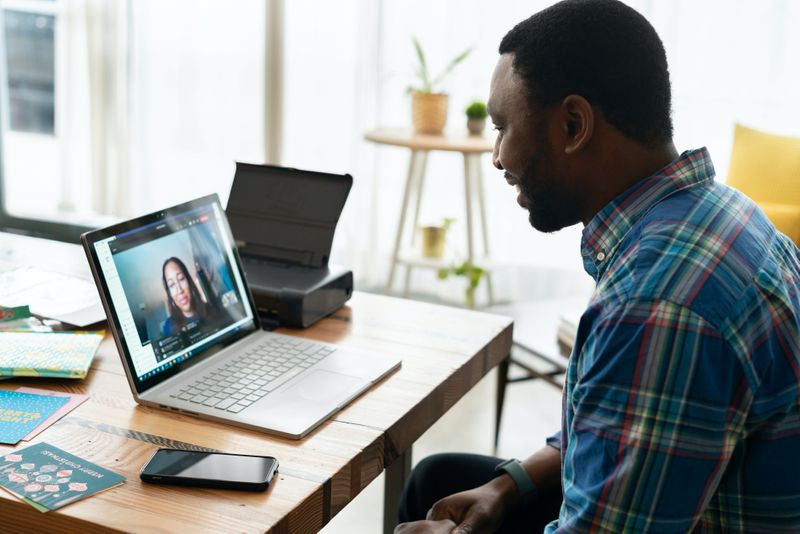 Two people talking.  A women is present in a laptop video conferencing, while the man is present at the table.