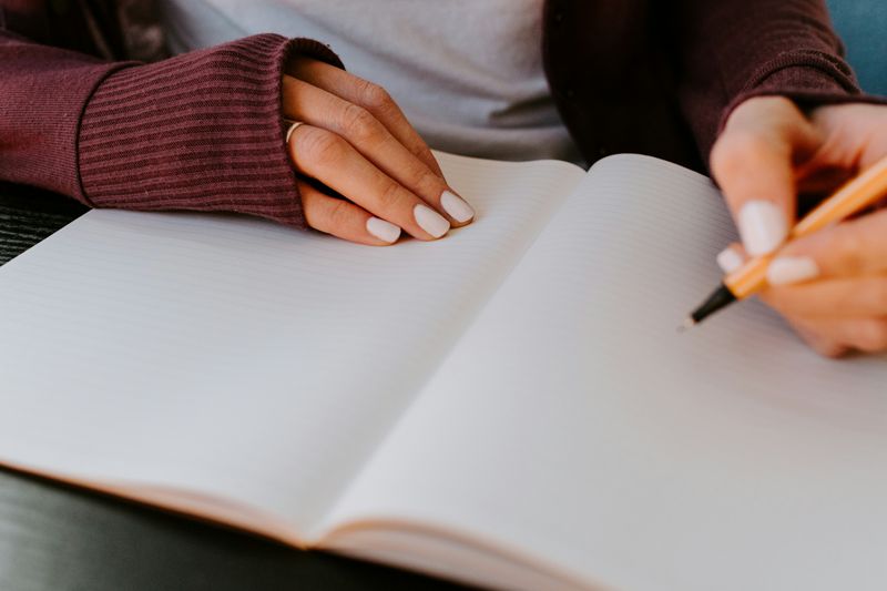 2 hands on a notebook. One hand holding down the book and the other is holding a pen beginning to write in the journal