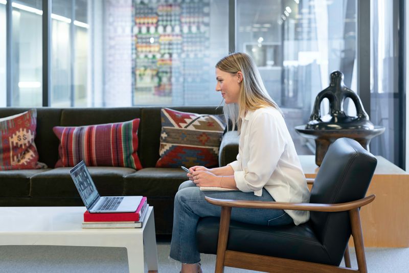 Image: Person with blonde hair wearing jeans sits in office building lounge for a virtual meeting with Western-printed pillow