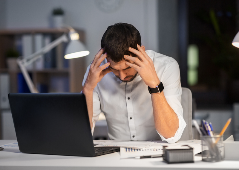 A man sitting at his desk, holding his head in his hands, looking stressed, with paperwork and a computer in front of him.