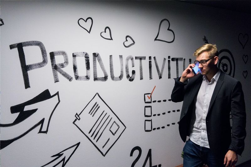 Image of a man holding a phone and standing in front of a wall that reads productivity. 