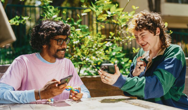 A man and a woman are smiling at each other while holding their phones, possibly sharing something pleasant.