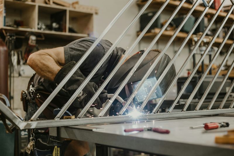 A welder leans over their work as they use a torch to join metal tubing together. They wear a face shield and thick gloves.