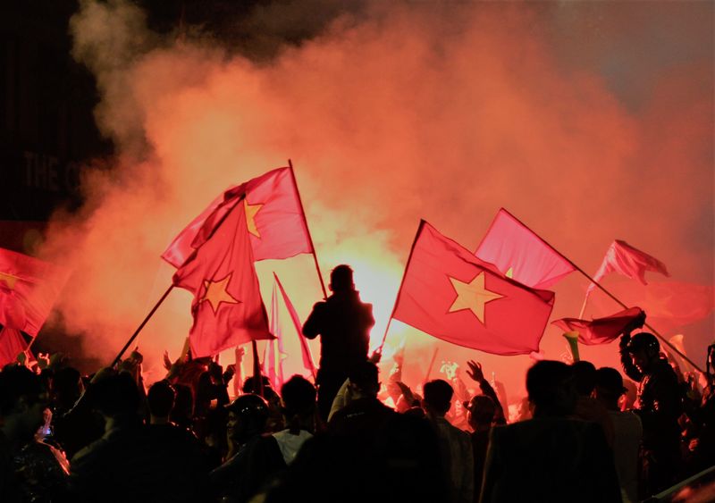 A rally in Vietnam. A group of people hold up the Vietnamese flag while flares go off in the background.