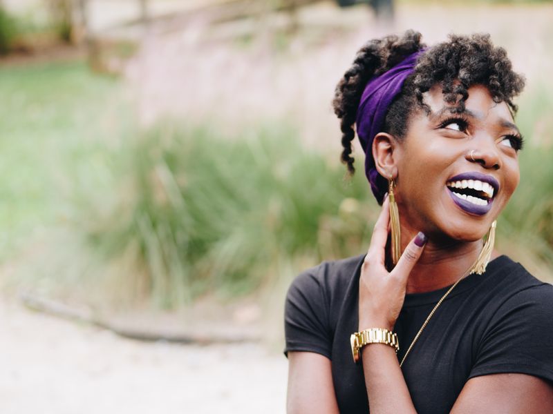 Woman wearing a purple headband and purple lipstick smiles and looks up to the right while her hand rests on her neck.