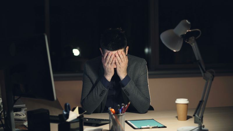 A man at his work desk covering his face with his hands.