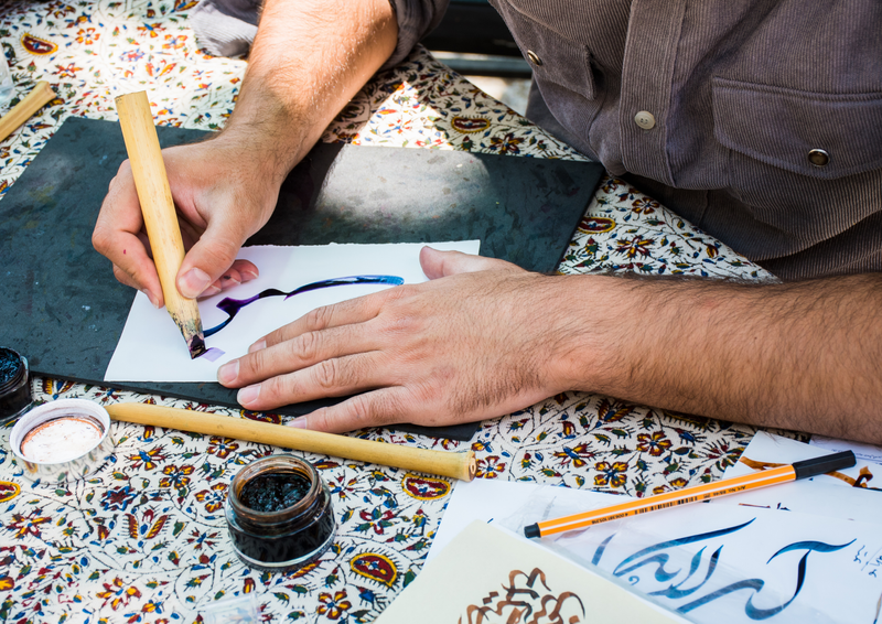 A man skillfully writing in Nastaliq, a traditional form of Persian calligraphy, using a reed pen and ink on white paper.