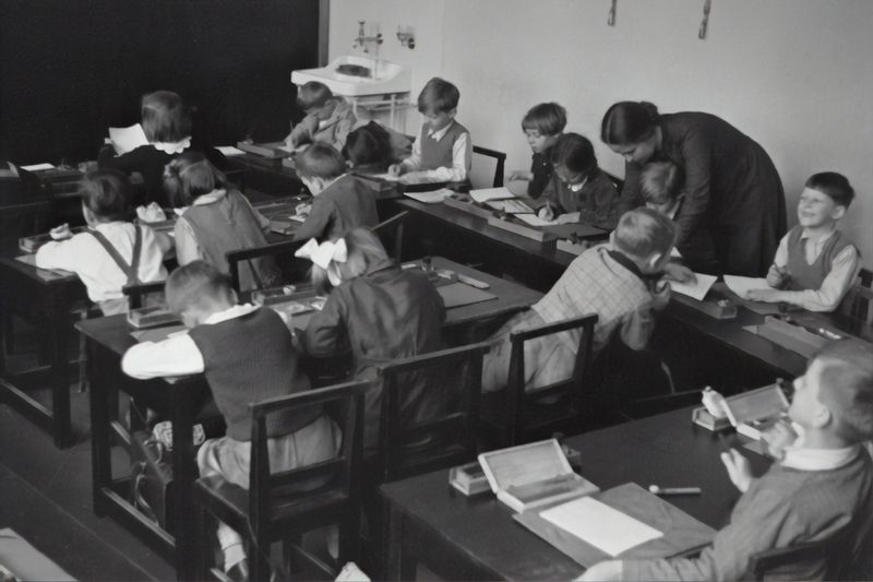 Historical black-and-white image of schoolchildren working at rows of desks while the teacher bends over to help a student.