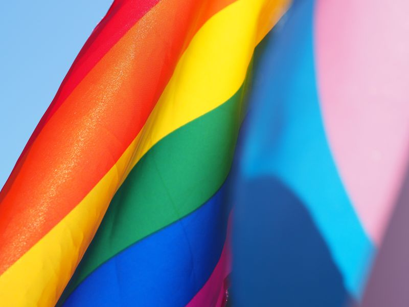 An LGBTQ+ flag and trans flag flying in front of a blue sky.