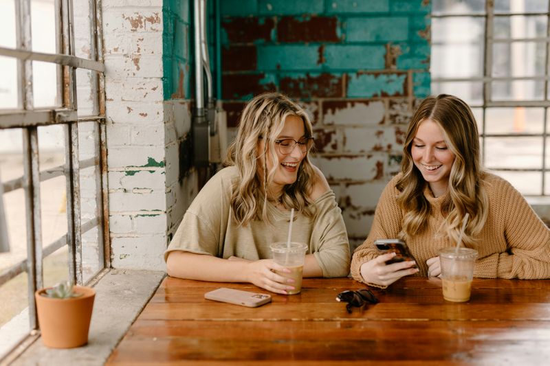 Two friends laughing while looking at a phone in a café.