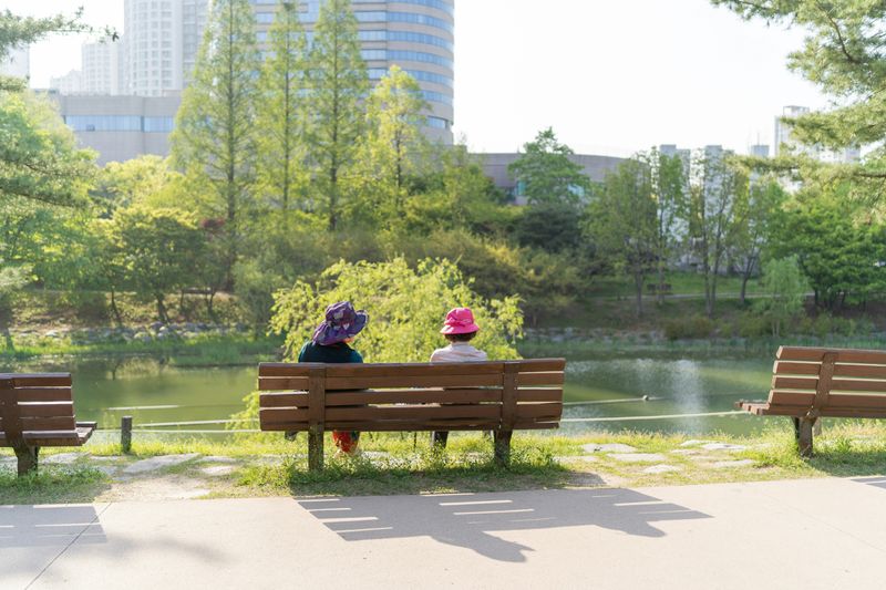 Two friends sitting on a bench near a pond outside.