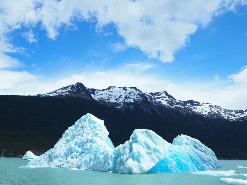 An iceberg floating in water with snow-capped peaks behind and clouds in the blue sky.