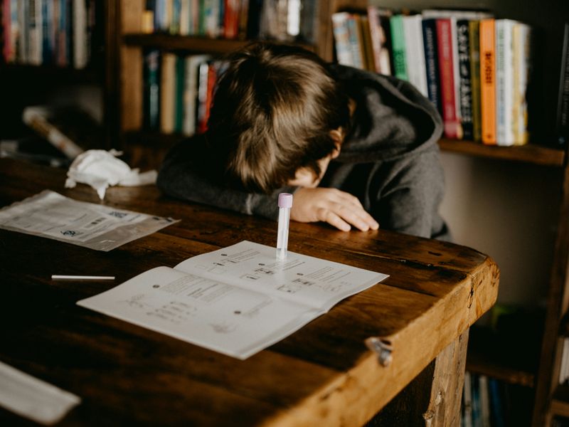 A student putting their head on a table, looking overwhelmed, with some school materials scattered around them.. 