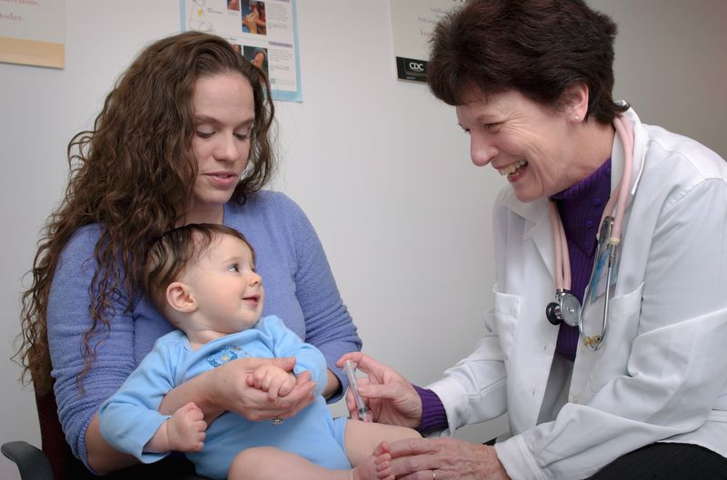 A pediatrician giving an injection to a baby, who sits in their mother's lap.
