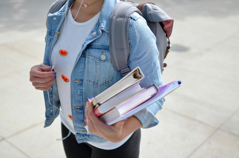 person wearing a light wash denim jacket holding books and wearing a backpack.