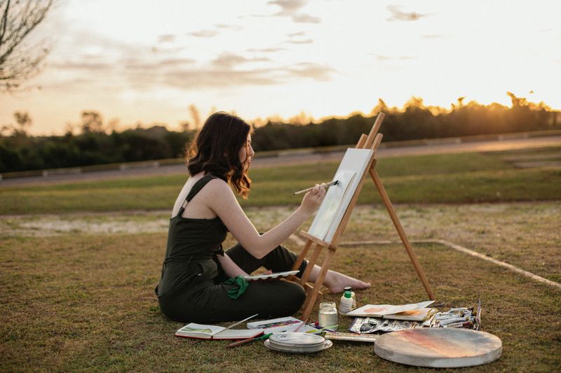 A person sitting in a grassy field in front of an easel, painting a nearby scene on a canvas.