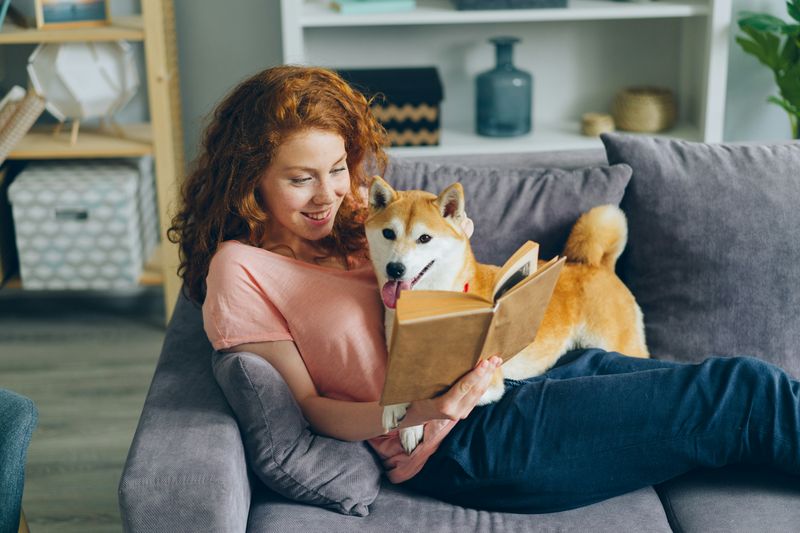 A woman sitting on the couch reading a book with her dog.