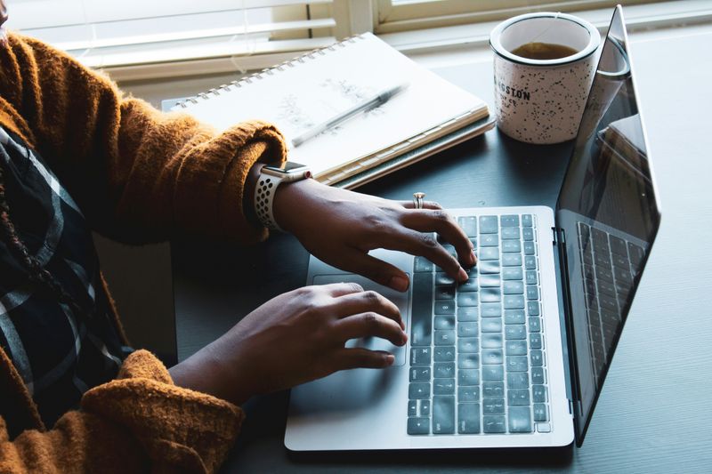 A person typing on a laptop at a desk with a notebook and mug beside them.