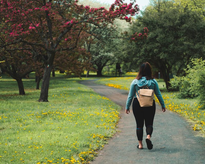 Woman with short brown hair in a casual outfit walking in a park with flowering trees and grass with yellow dandelions in it