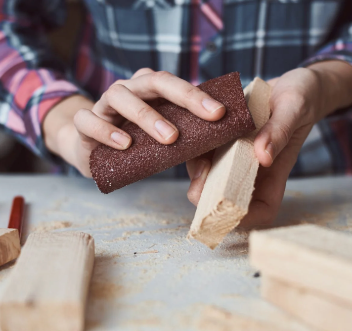 A person in a plaid shirt holding a piece of red sandpaper and rubbing it against a block of pale wood.
