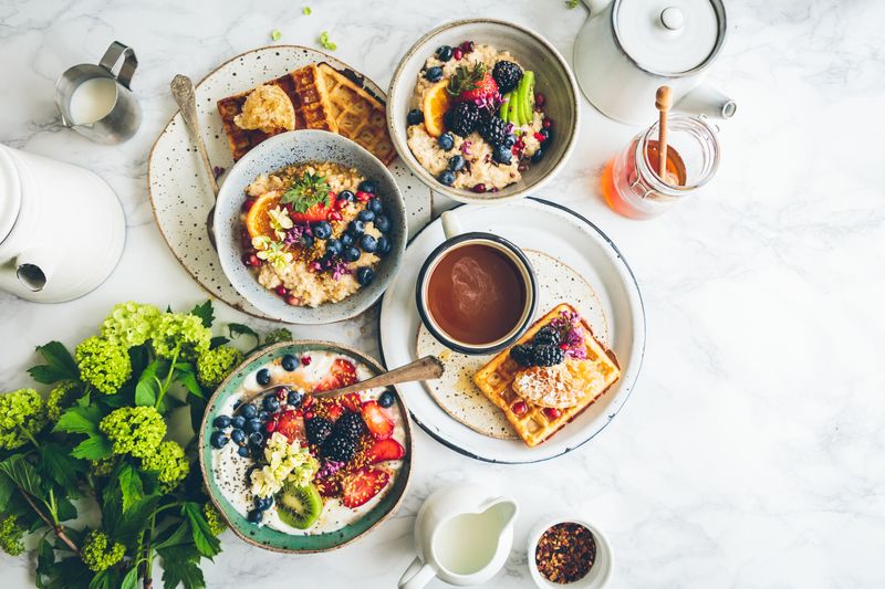 A table with several plates of breakfast. On the plates are waffles, fruits, berries, and oatmeal.