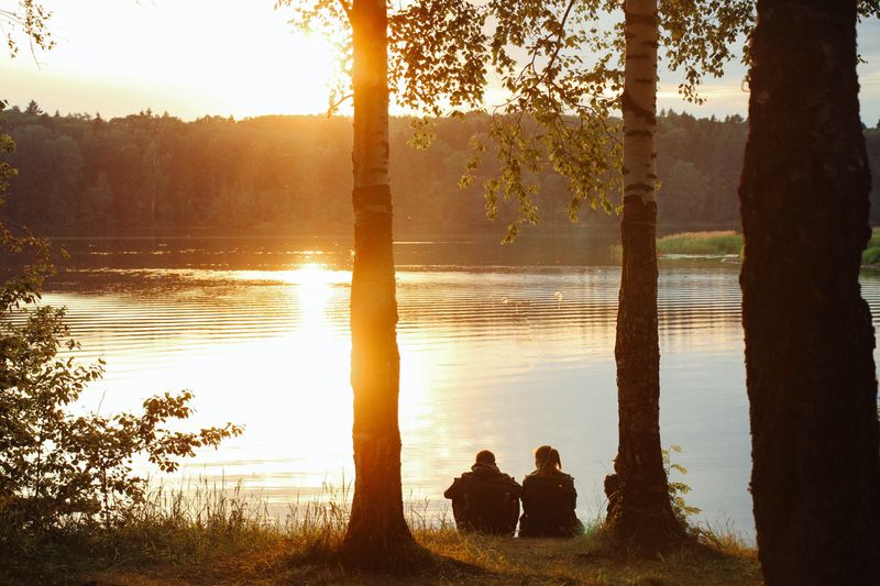 Two people sitting at the edge of a lake, facing a sunset.