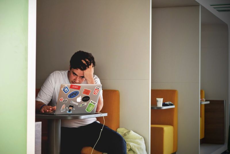 A stressed person sits at a booth in a cafe looking at a laptop