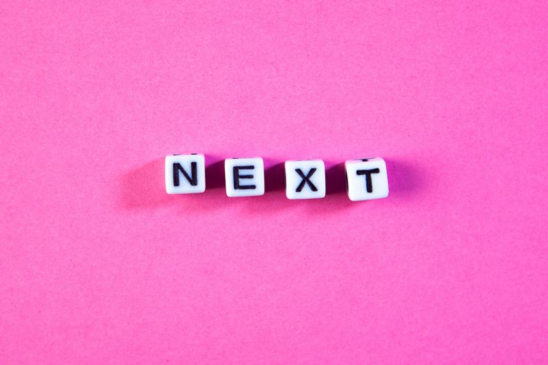 The word "NEXT" spelled out with white letter beads centered on a bright pink background.