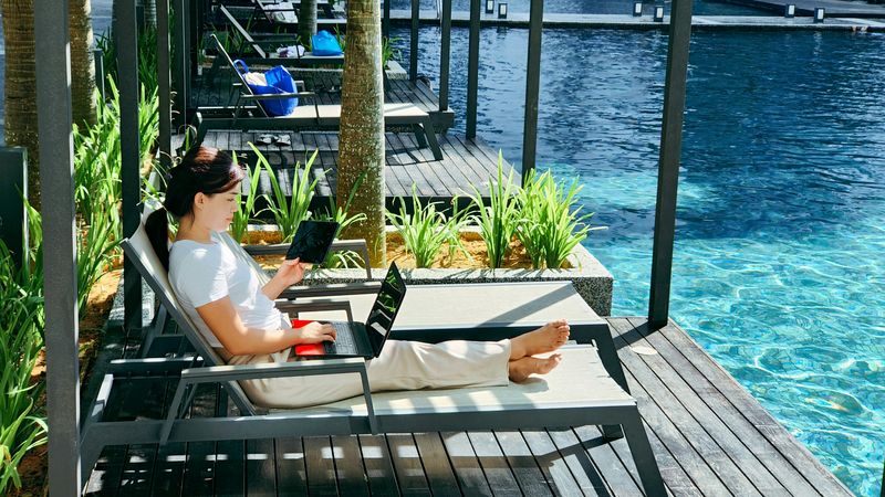 A digital nomad sitting in a lounge chair with their laptop by a hotel pool.