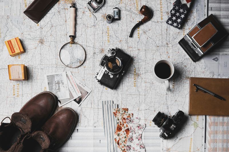 A travel-themed flat lay featuring a map, vintage cameras, a magnifying glass, a pipe, a notebook, coffee, and boots.