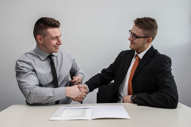 Two men in business clothing sit behind a table, shaking hands over papers.