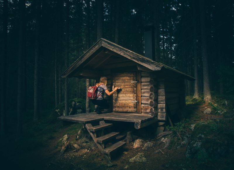 Deep in the woods, a woman with a backpack kneels to enter a log cabin the size of a sleeping tent.