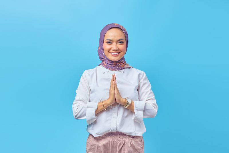 Smiling woman with purple hijab and praying hands stands in front of blue background