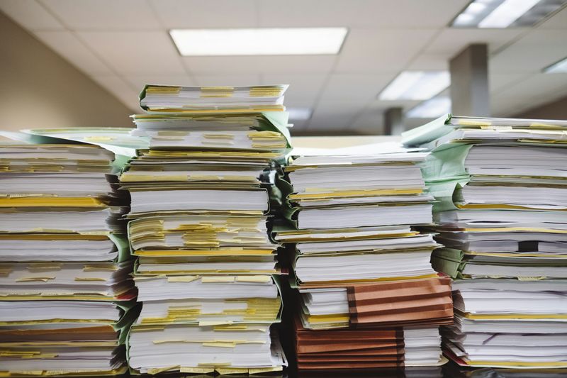 Tall stacks of paper documents and folders filled with yellow sticky tabs sitting on an office desk.