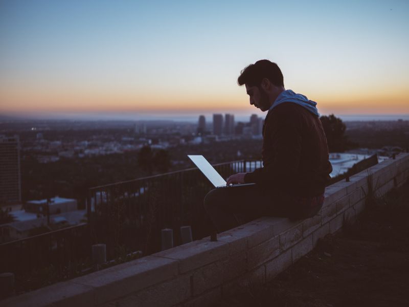 A person sitting on the edge of a rooftop while working on a laptop.