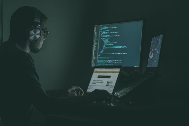 Man with headphones and glasses sitting in dark room in front of three computer monitors