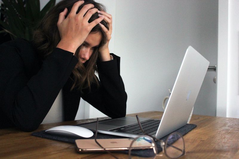 Woman has hands on her head as she stares at her laptop in frustration. Her glasses sit on the table.