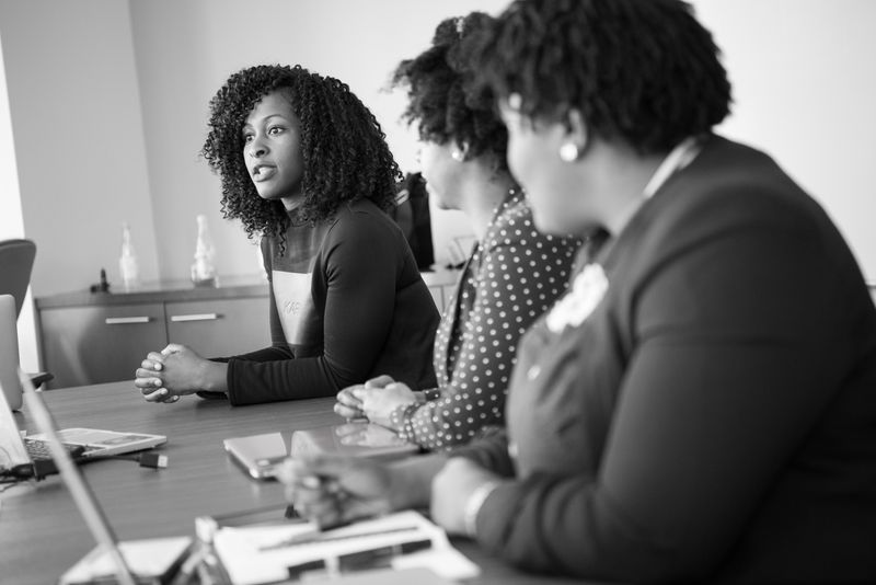 A young professional woman with black curly hair confidently speaks in a meeting room of other women